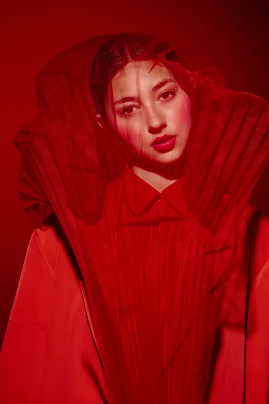 Fashion woman in red with dramatic translucent veil covering face gazes softly, studio portrait captures bold beauty and creative styling, beauty and fashion concept.