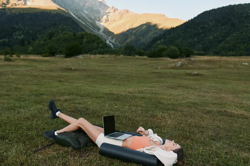 A person lies on a grassy field with a laptop on a mat, surrounded by mountains. The scene is peaceful yet productive, blending nature with remote work.
