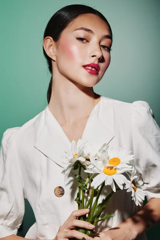Beautiful young woman with flawless makeup and glossy lips holding fresh daisies wearing a white buttoned dress against green background. Concepts of beauty and fashion.