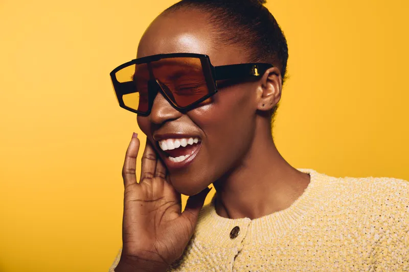 Happy African woman with dark skin wearing oversized sunglasses and a yellow sweater smiles with closed eyes on yellow background. Concept of fashion, joy, and beauty portrait.