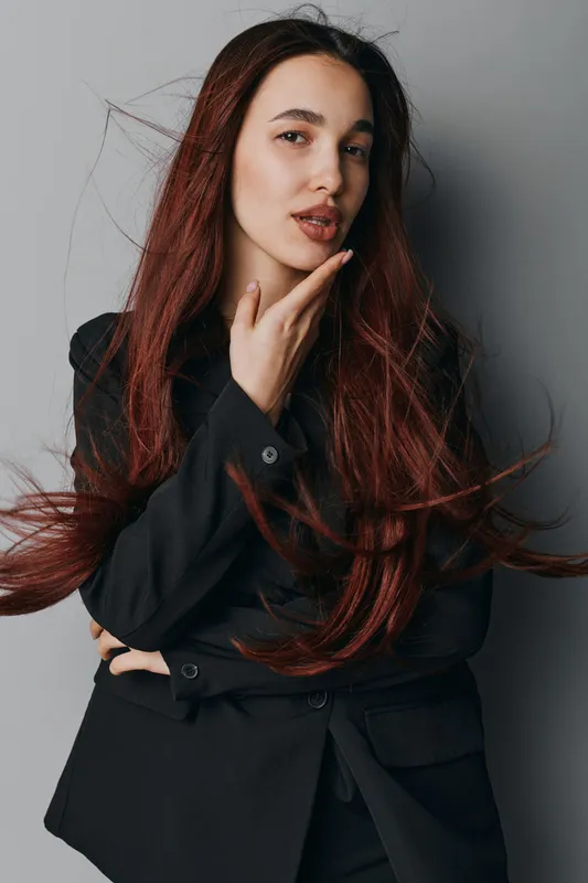 Woman with long red hair wearing black blazer posing with hand on chin and looking at camera on gray background. Studio portrait of confident female with natural makeup and flowing hair.
