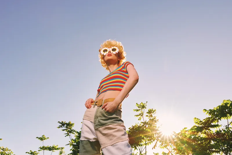 A young woman wearing a striped shirt and flower crown stands outdoors as the sun glints, conveying a carefree retro vibe and summer nostalgia.