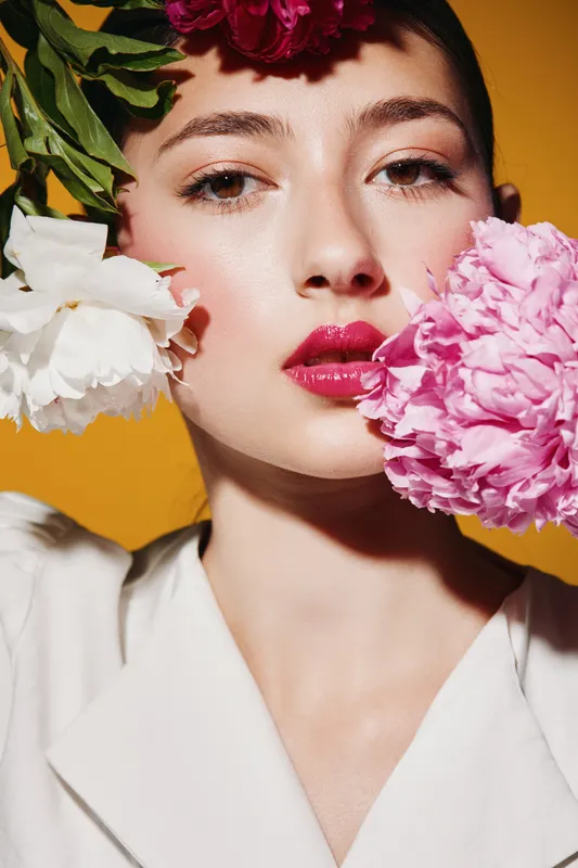 Close-up portrait of a young woman with pink and white flowers near her face against an orange background. Beauty fashion creative shoot with vibrant makeup and fresh petals.