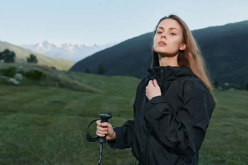 A young woman stands in a grassy alpine meadow, wearing a dark windbreaker and gripping a trekking pole. She gazes ahead with a calm, confident expression against distant mountains.