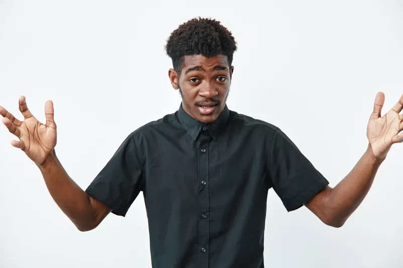 Young black man with dark skin wearing black shirt standing with arms open and expressive face on white background. Casual style and confident gesture in studio.
