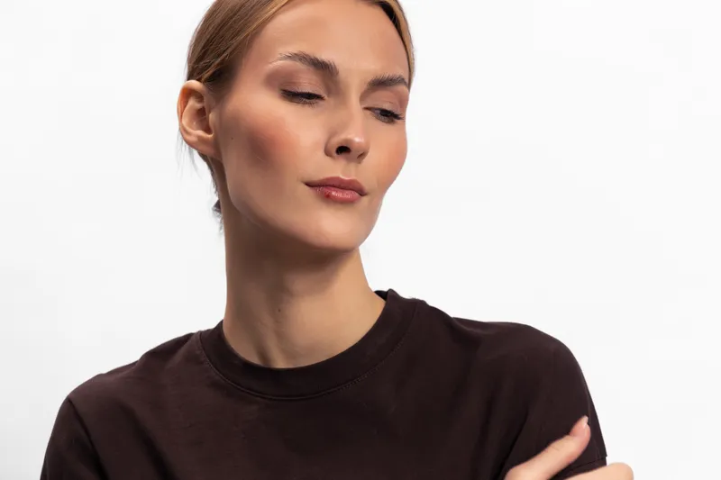 Calm young woman with light skin and closed eyes wearing a dark brown shirt poses against white background. Concept of serenity, confidence, and natural beauty with soft lighting.