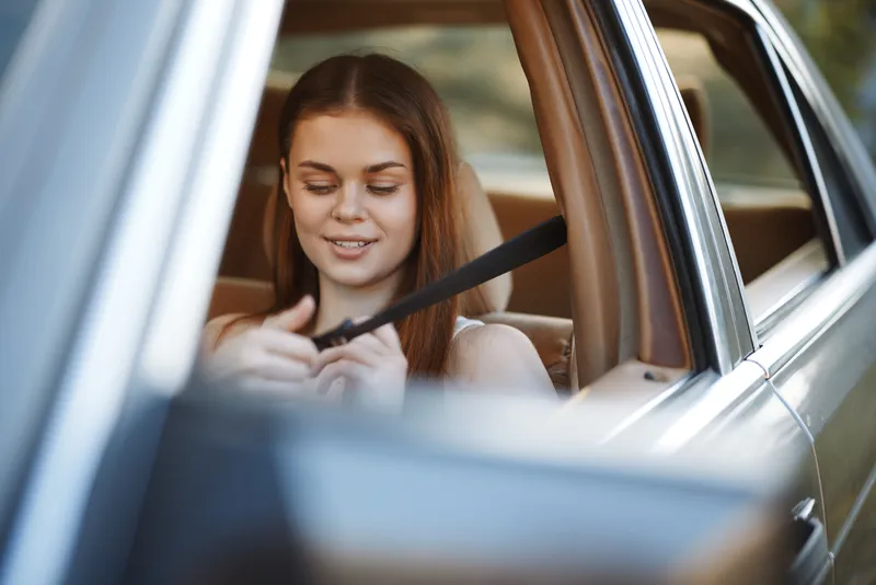 Young woman fastening her seatbelt inside a car, showcasing safety and comfort while enjoying a sunny drive, with a focus on her joyful expression and casual attire.