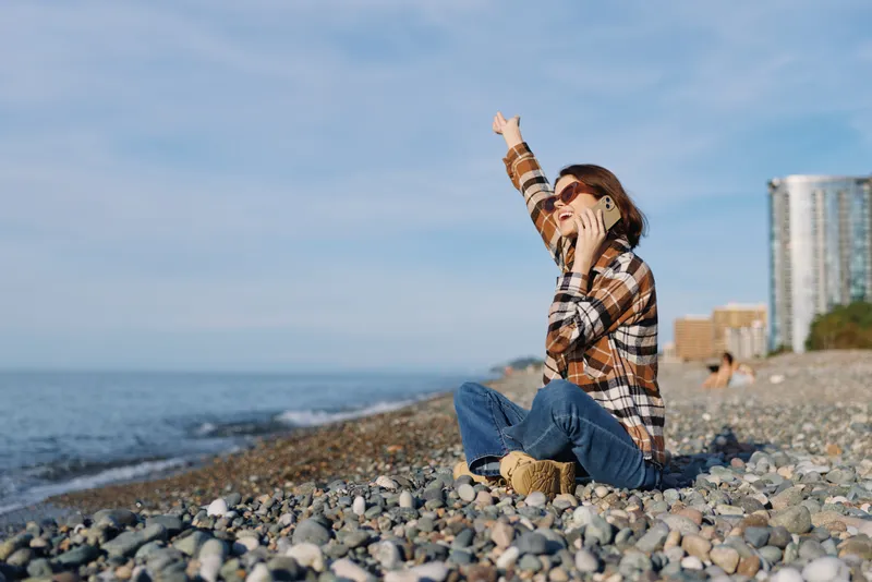 Female traveler on a pebbled beach talking on a phone, waving toward the horizon with calm sea, distant skyline and clear blue sky creating a relaxed urban seaside moment