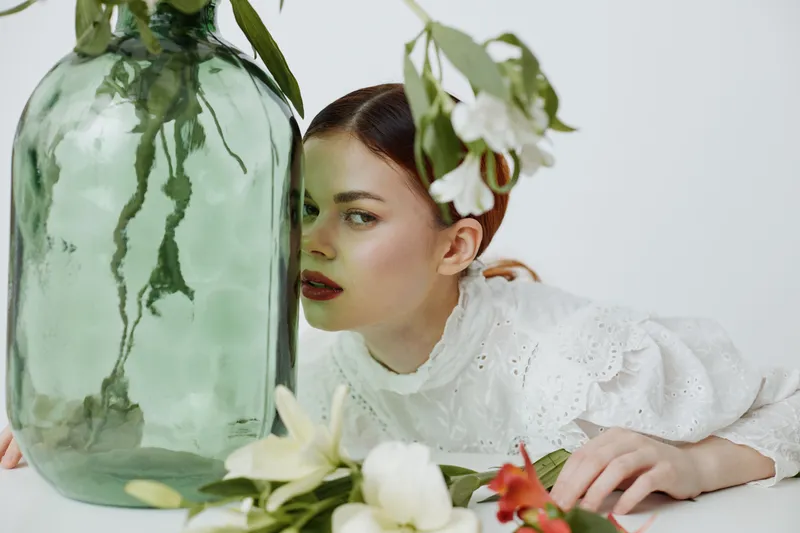 Young woman with pale skin and red hair, dressed in a white vintage blouse, gazing through a green glass vase surrounded by fresh flowers