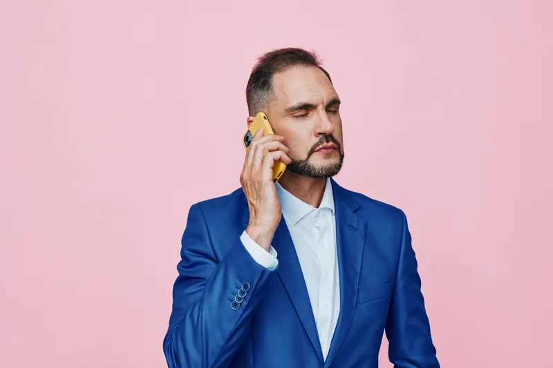 A polished business professional talks on a mobile phone against a solid pink backdrop, conveying confidence, focus, and calm as he handles a strategic moment.