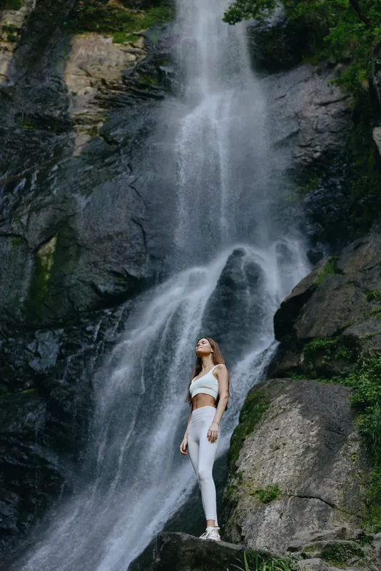 Young woman standing near powerful waterfall in nature, dressed in white activewear, surrounded by rocks and greenery, symbolizing outdoor adventure and fitness.