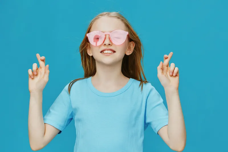 Happy girl with pink sunglasses and crossed fingers on a bright blue background, expressing joy and optimism with a playful summer vibe