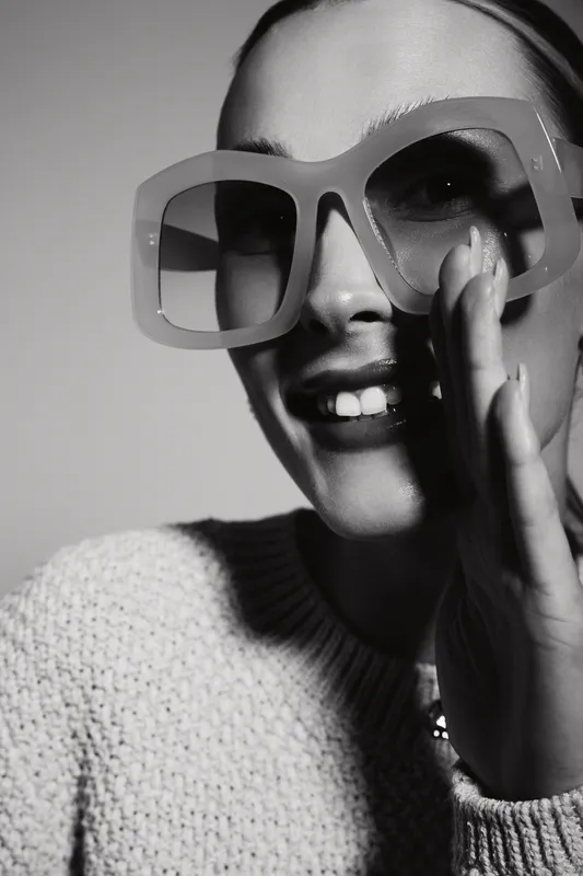 Stylish caucasian woman with light skin wearing large sunglasses and a knitted sweater smiles, whispering with hand near mouth in black and white portrait. Concept of fashion and communication.