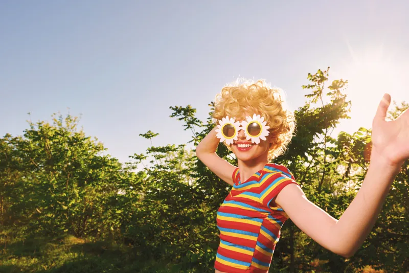 Smiling woman with curly hair wearing a striped top in a sunny outdoor setting, retro color grading, bright sunglasses with flower shapes, joyful lifestyle moment in nature.