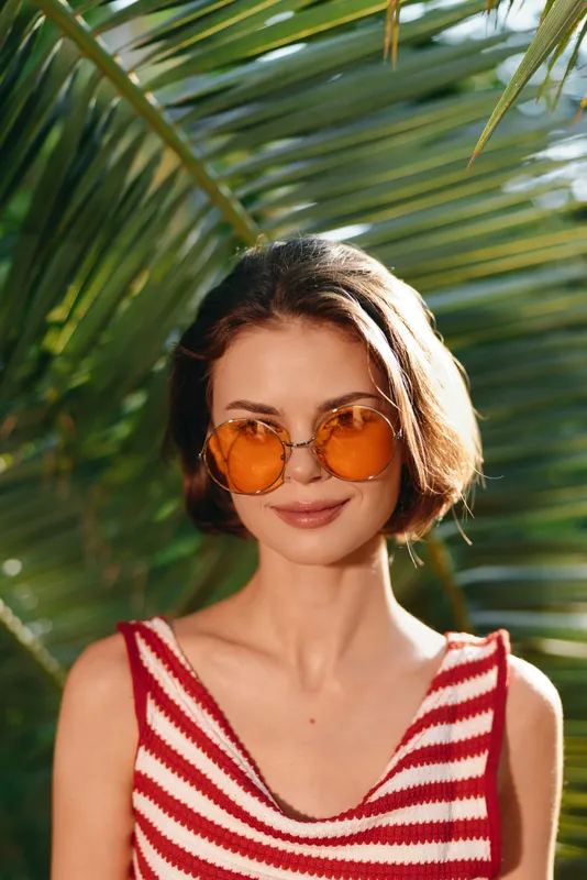 An elegant woman wearing a striped red and white top stands outdoors among lush tropical plants, smiling softly, radiating casual confidence and a bright summer vibe under warm light.