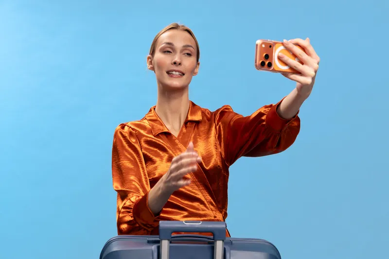 Young woman in orange blouse takes a selfie with smartphone while holding suitcase handle on blue background. Concept of travel, vacation, tourism, and modern lifestyle.