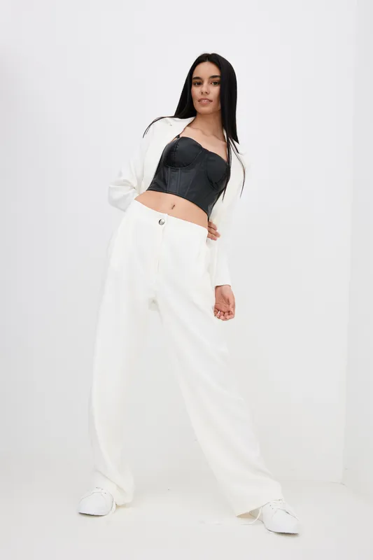 young woman dressed in white pants and black corset blouse posing confidently against plain white backdrop, showcasing modern fashion style in studio setting