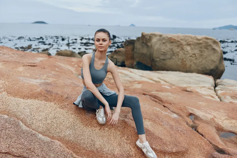 Young woman in activewear practicing yoga on rocky beach, promoting wellness and healthy lifestyle with serene ocean background and soft color palette