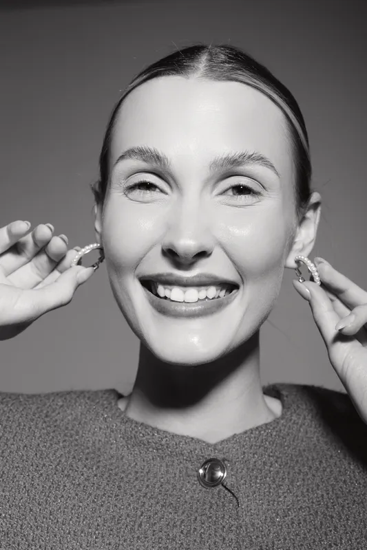 Portrait of a smiling white woman with fair skin holding earrings near her face, studio lighting on grey background.