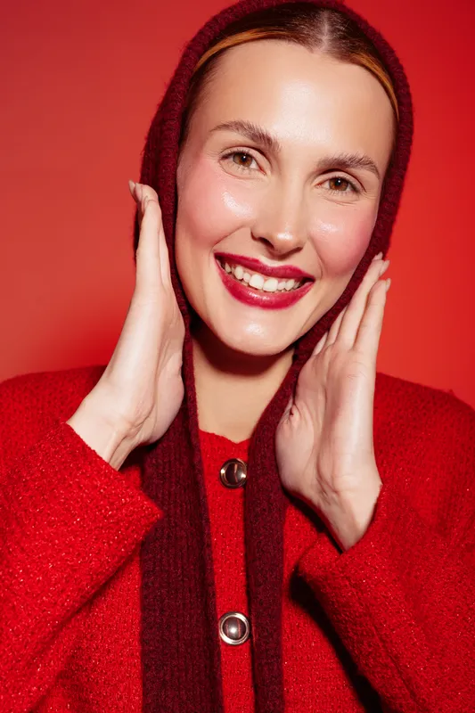 Smiling woman in red sweater and scarf poses against red background. Close-up portrait with natural makeup and bright lipstick. Concept of fashion, beauty, and winter style.