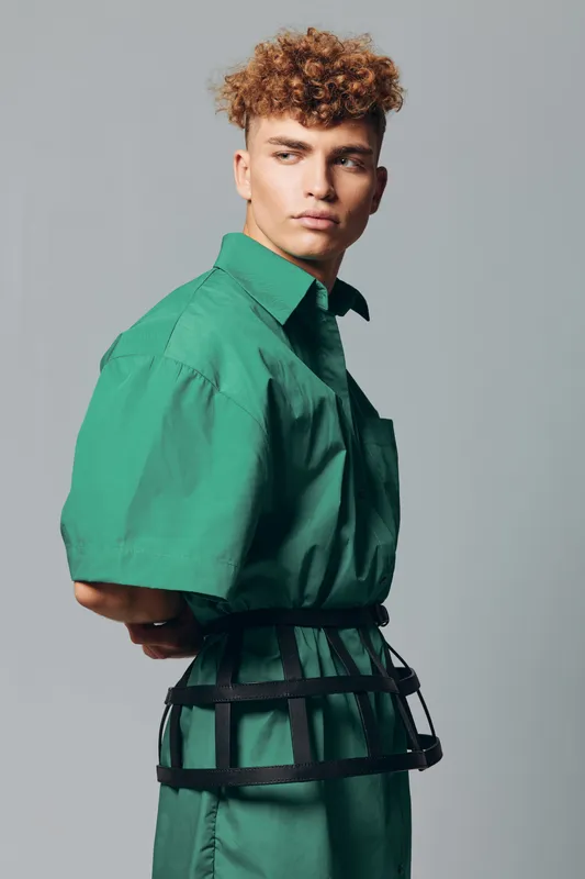 Young man with curly hair wearing green shirt and black harness posing sideways on gray background. Fashion model with serious expression and modern hairstyle in studio.