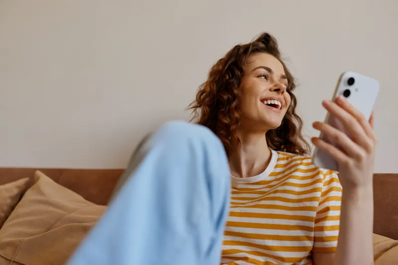 Woman enjoys a relaxed moment at home, smiling as she uses a smartphone, seated on a couch in a striped tee and jeans, capturing casual daily life with warm natural light.