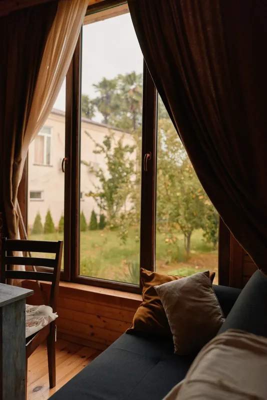 Cozy living room scene with a large window, warm sunlight, plush sofa and cushions, and flowing curtains creating a relaxing and inviting home atmosphere.