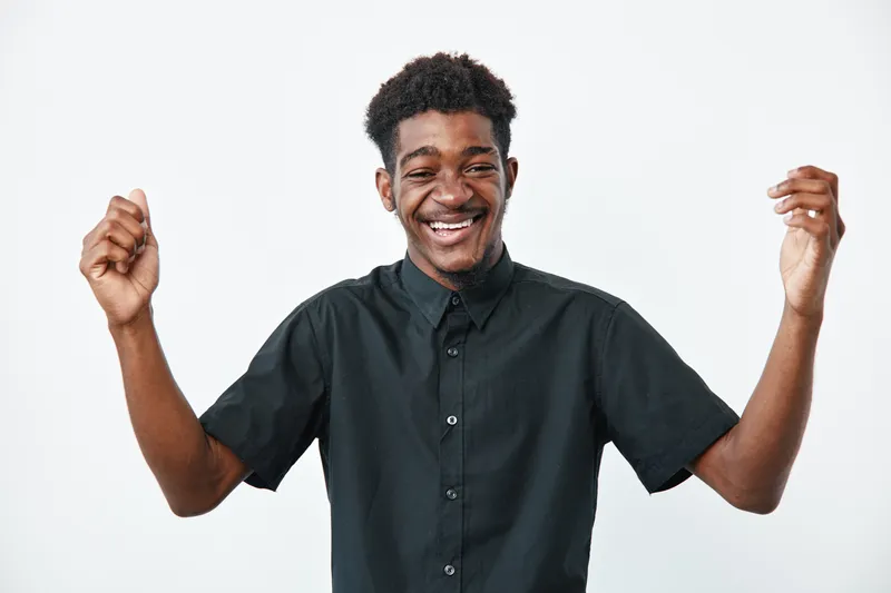 Young black man with dark skin wearing black shirt smiling and raising hands in celebration isolated on white background. Happy African male expressing joy and success.