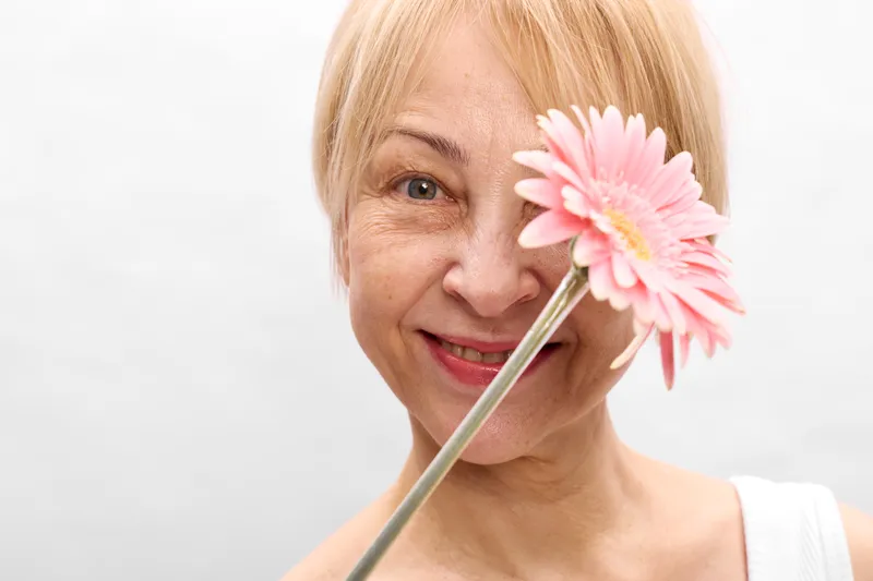 Woman smiling older holds flower near face, happiness in spring portrait, closeup on white background, pink gerbera beside cheek with soft light, calm joy