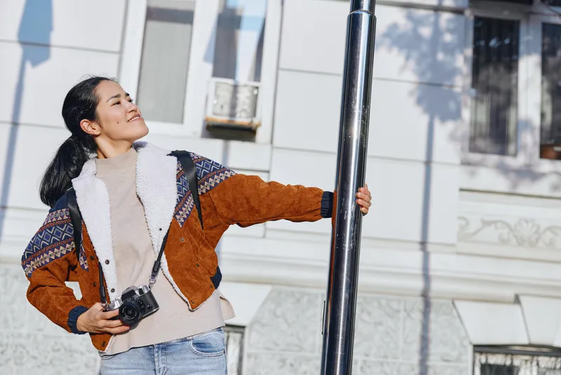 Woman holding a camera outdoors near a lamppost, smiling toward daylight, wearing a brown shearling coat and cozy sweater, casual street style captured in bright urban light and breeze