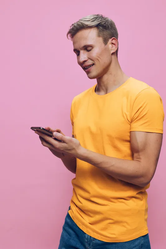 Man in casual yellow t-shirt holding and looking at mobile phone isolated on pink background studio portrait. People lifestyle concept