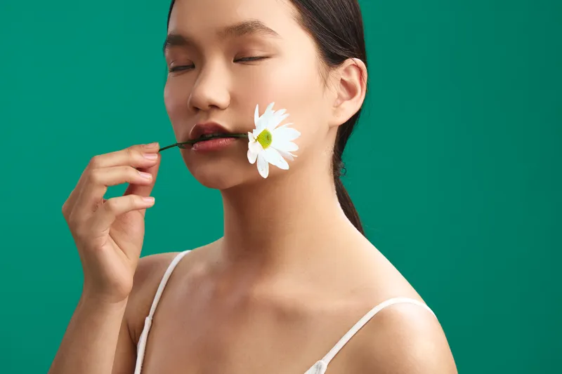 Asian woman with a delicate white daisy held at the lips, wearing a light tank top. She stands against a vivid green backdrop, exuding calm elegance and subtle artistry in a refined studio setting.
