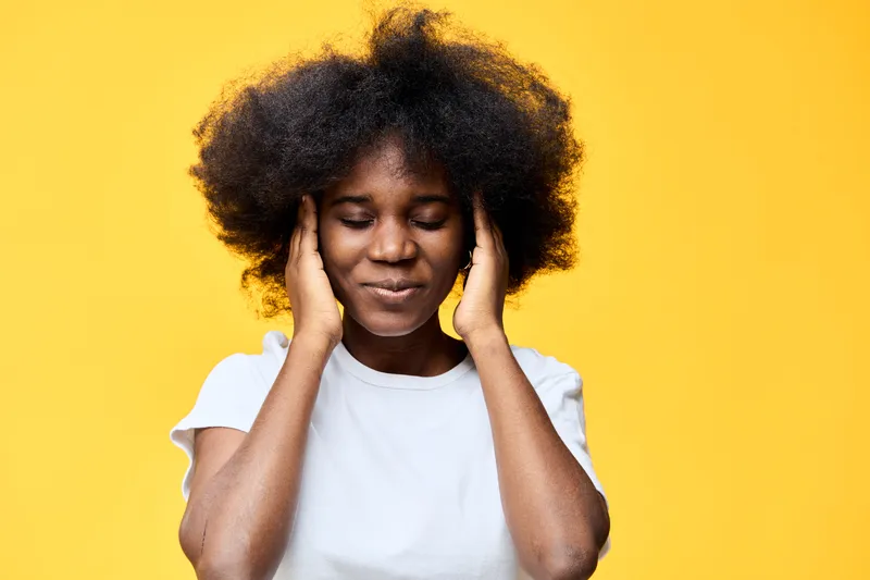 Young African woman with curly hair smiling while covering her ears, against a vibrant yellow background, conveying joy and tranquility in a cheerful setting