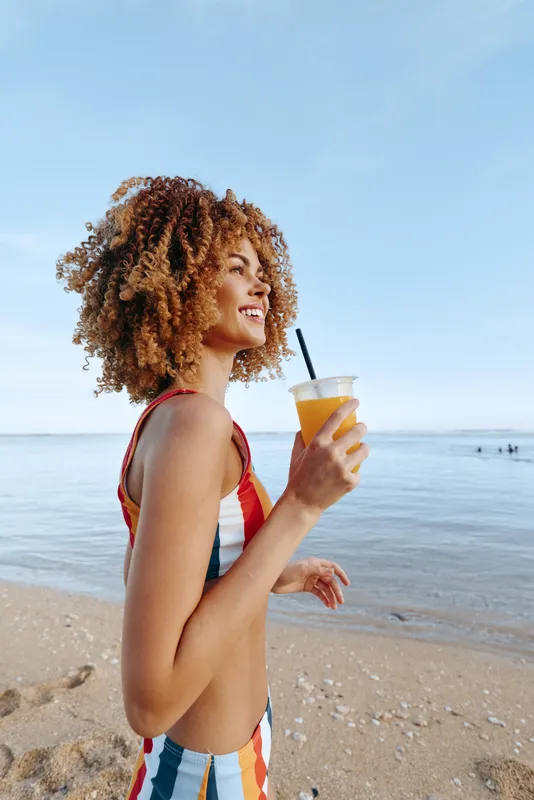 Young woman in striped swimsuit walks along a sunny beach, sipping juice from a cup and smiling toward the sea, with a gentle breeze and golden sand under clear skies.