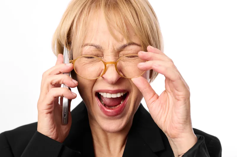 Laughing business woman in suit, eyes closed, adjusting glasses, holding smartphone at ear, joyful moment, studio white background, soft light, close up