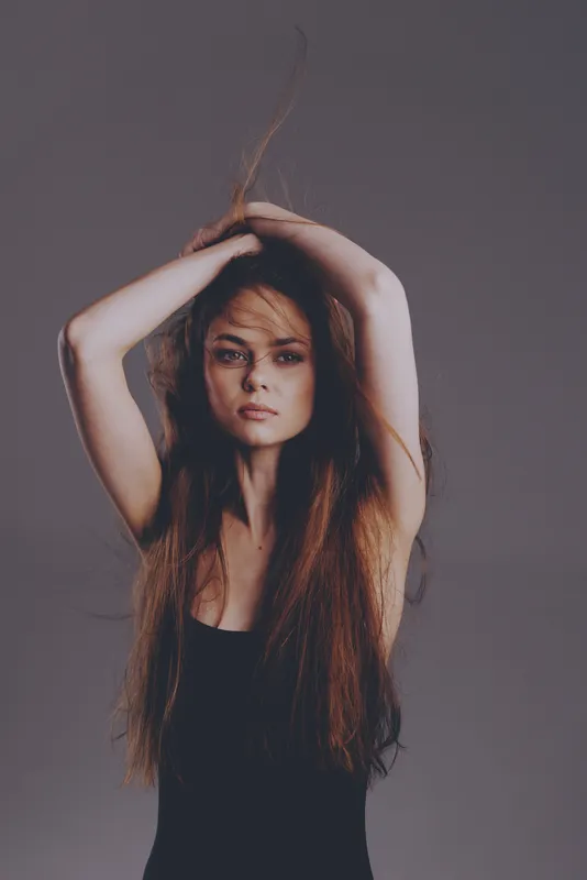 Serious woman with long hair posing with raised arms against gray background in studio lighting.