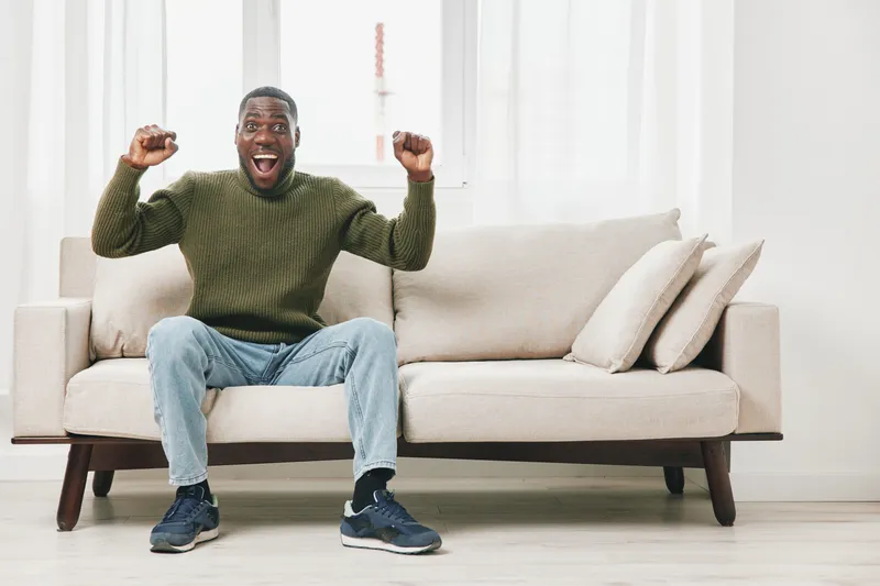 Excited male model in a green sweater celebrating on a couch, showcasing joy and enthusiasm in a modern, bright living room setting