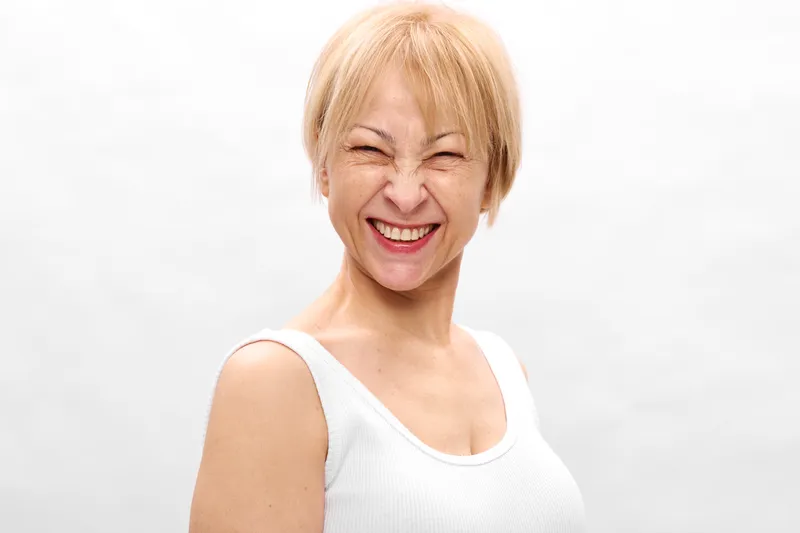Smiling woman, elderly, blonde hair, white top, close portrait on white background, studio lighting, wellness mood, cheerful expression, soft light, indoor calm