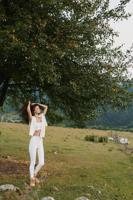 Young woman in white outfit stands in an open field beneath a leafy tree, striking a casual pose with lifted arms and a relaxed joyful mood