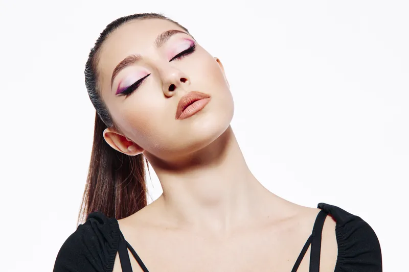 Young woman with pastel pink winged eyeliner and nude lipstick poses with closed eyes on a white background, wearing a black outfit. Concept for beauty, fashion, and makeup artistry.