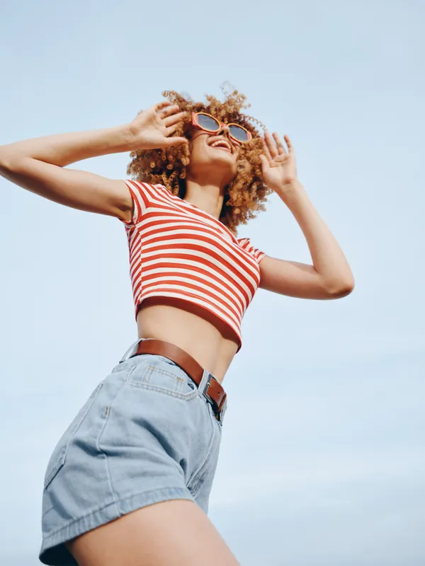 A carefree young woman wearing a red striped cropped top and denim shorts raises her arms against a bright blue sky, capturing a vibrant, playful summer moment.
