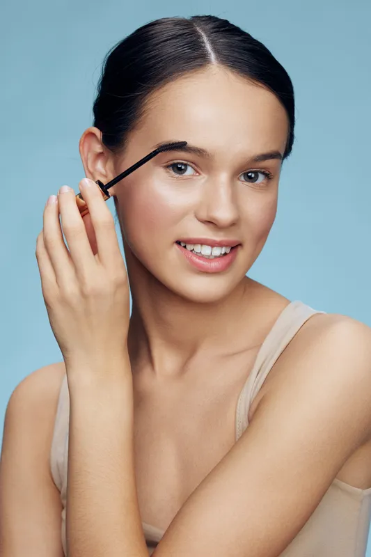 Young woman applying mascara with a joyful expression against a soft blue background, showcasing beauty and self care in modern makeup