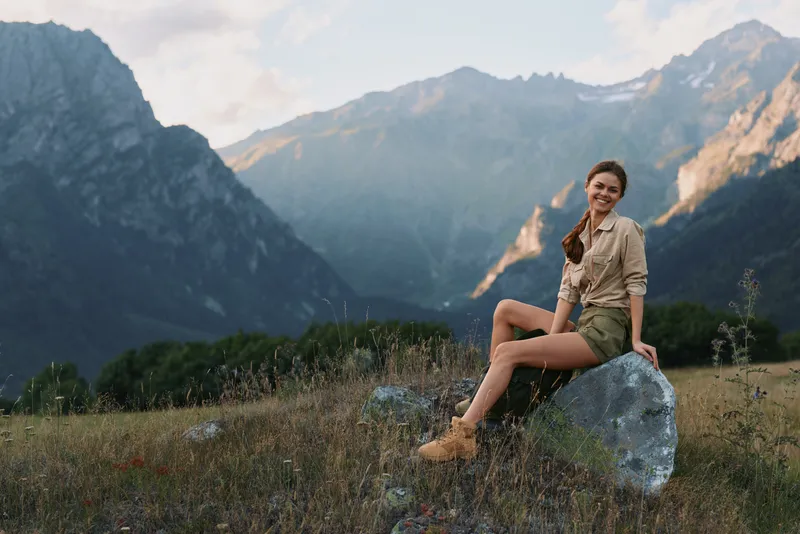 A young woman in casual clothes sits on a rock in a mountain meadow, smiling and enjoying nature during a sunny day. Concept for lifestyle, travel, and outdoor relaxation.