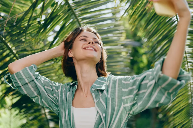 A cheerful woman outdoors with arms raised under sunny palm leaves, enjoying a carefree moment in bright daylight.