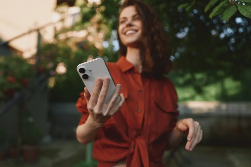 Woman lifestyle outdoor selfie in daytime, smiling, wearing red shirt, casual fashion, holding smartphone, cheerful candid portrait in garden setting with natural light and warmth