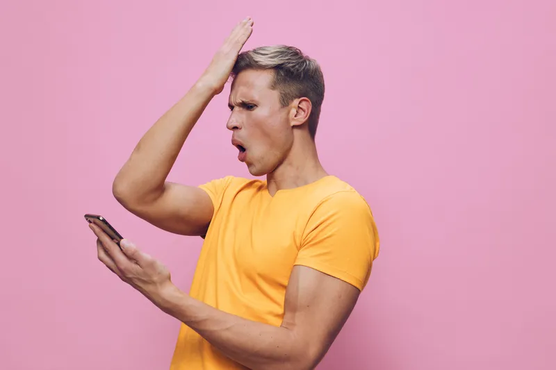 frustrated young man wearing yellow tshirt holding smartphone looking shocked surprised isolated pink background digital detox concept