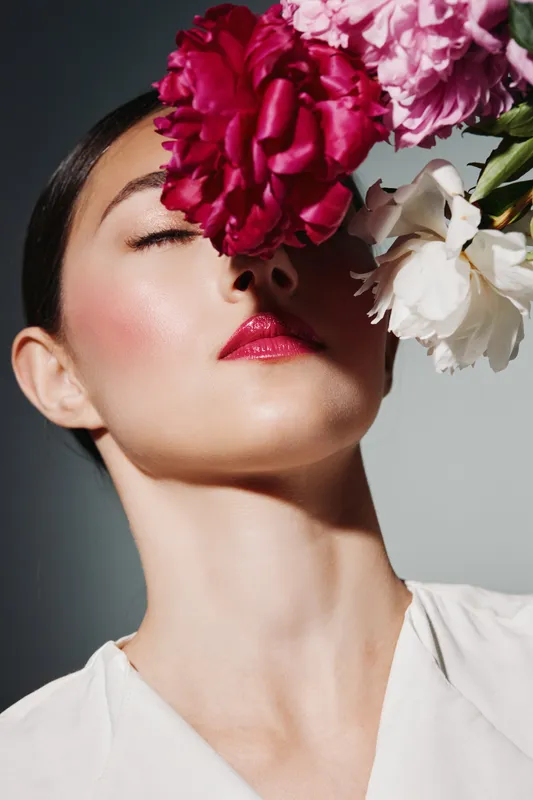 Fashion model with flawless glowing skin and shiny red lips poses with face partially covered by large bright flowers. Creative beauty shot with soft natural lighting.