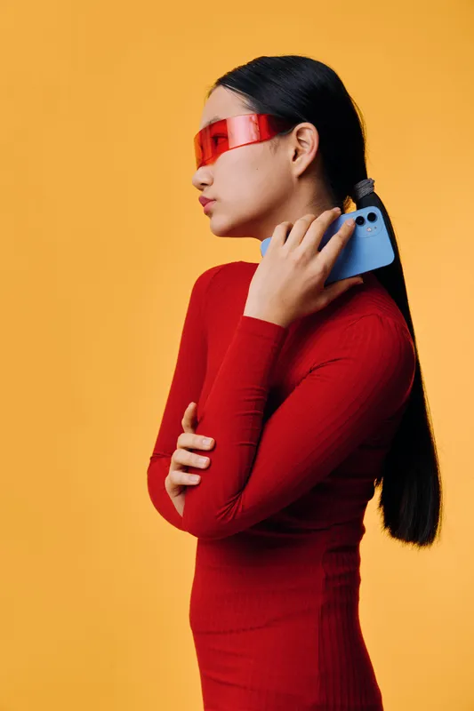 Young woman in red dress wearing futuristic red visor holding a blue smartphone over shoulder on orange background with long black hair and confident pose in modern style fashion.