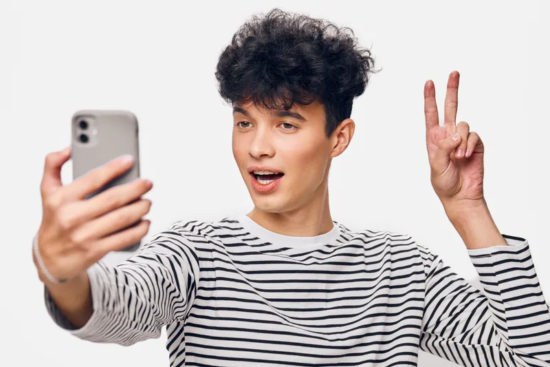 young man taking selfie with smartphone showing peace sign, casual striped shirt, cheerful expression, isolated white background, modern lifestyle