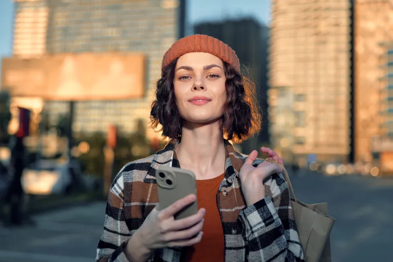 Woman with smartphone in urban street portrait, candid lifestyle moment at golden hour glow, beanie and shopping bag, authenticity and mindful living captured with emotional storytelling.